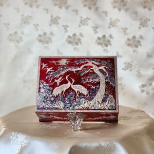 May include: A red wooden box with a hinged lid decorated with a mother-of-pearl inlay of two cranes standing under a pine tree. The box has a silver butterfly-shaped clasp.