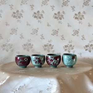 May include: Four small ceramic teacups with floral designs in shades of burgundy and white on a light blue background. The cups are arranged in a row on a white tablecloth.