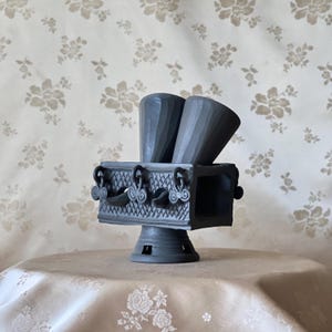 Può includere: Un bruciatore di incenso in ceramica grigia con due supporti a forma di cono e una base rettangolare. La base presenta manici decorativi e un piedistallo. Lo sfondo è un motivo floreale.