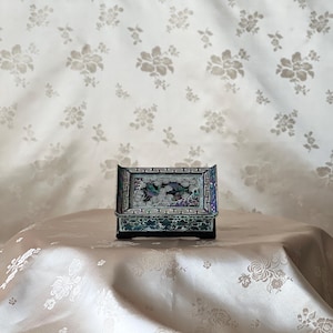 May include: A small rectangular black box featuring intricate mother-of-pearl inlay on the lid and sides. The lid displays a detailed scene of birds amidst floral patterns, framed by a geometric border, with additional floral motifs on the base.