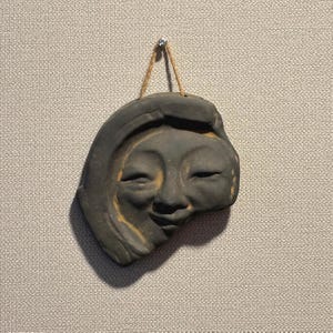 May include: A dark gray ceramic wall hanging depicting a smiling face with closed eyes. The sculpture is suspended by a natural fiber cord. The face has a serene expression and is mounted on a textured, light gray wall.
