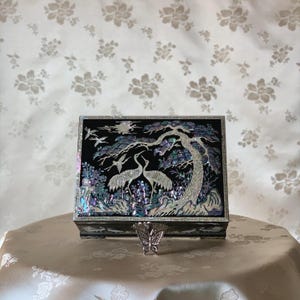 May include: A black jewelry box with a mother-of-pearl inlay design featuring two cranes under a tree. The box has a silver butterfly-shaped stand in front of it. The background is a floral patterned fabric.