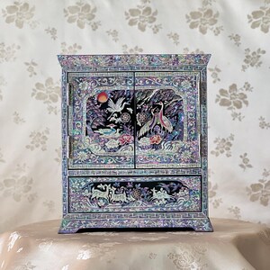 May include: An iridescent jewellery box with a rectangular shape. The box features intricate mother-of-pearl inlay with images of birds, flowers, and deer. The box has two doors and a drawer. It sits on a table.