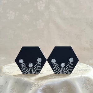 May include: Two black hexagonal coasters with white floral designs. The coasters have a simple, modern design and are perfect for adding a touch of elegance to any home.