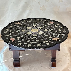 Puede incluir: Una mesa baja de madera oscura con borde festoneado. La tapa negra está incrustada con mariposas y diseños florales de nácar. La mesa tiene cuatro patas cortas y cuadradas.