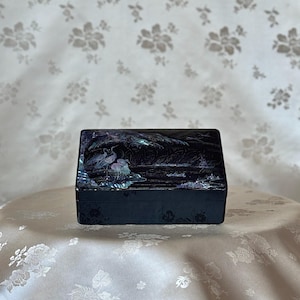 May include: A black wooden box with a hinged lid decorated with a mother-of-pearl inlay depicting a scene of birds and trees. The box is approximately 6 inches long and 4 inches wide.