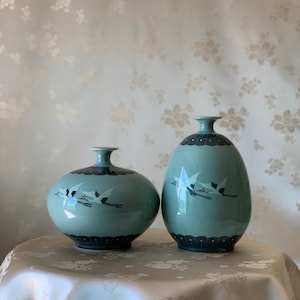May include: Two ceramic vases with a light blue glaze and black bird designs. The larger vase is oval shaped and the smaller vase is round. Both vases have a black rim.