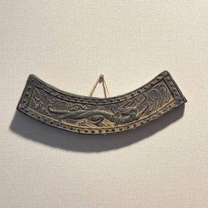 Può includere: Un pezzo decorativo in metallo scuro, curvo, con intricate incisioni a rilievo. Il design presenta figure di animali stilizzate e motivi floreali. Il pezzo è sospeso da una corda, possibilmente un manufatto storico.