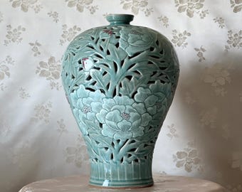 May include: A celadon-colored ceramic vase with intricate cut-out floral designs. The vase features large peony blossoms and foliage, with a detailed, openwork pattern. The vase is displayed on a table with a floral patterned tablecloth.