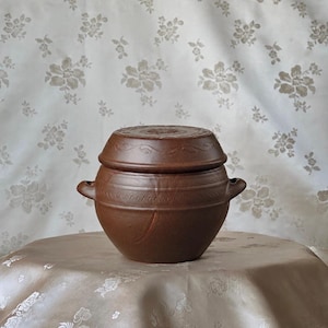 May include: A brown, round, lidded ceramic pot with two small side handles. The pot features a textured surface with a horizontal band of incised geometric patterns and a matching lid with a central floral motif and a delicate leaf border design.