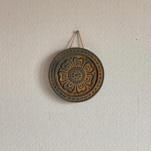 May include: A round, dark gray wall hanging with a floral design in the center. The flower has a light brown color. The outer edge has a series of small, raised dots. A natural fiber cord is used for hanging.