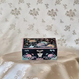 May include: A black wooden box with a hinged lid decorated with a floral and butterfly design made of mother of pearl. The box has a small metal latch.