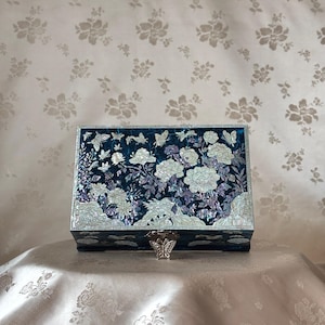 Könnte beinhalten: Eine rechteckige, dunkelblaue Box mit einem dekorativen Blumen- und Schmetterlingsdesign. Die Box ist mit schillernden Perlmutt-Inlays verziert, die weiße Blumen und Schmetterlinge zeigen. Ein silberfarbener Verschluss ist vorne sichtbar.