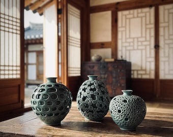 May include: Three green ceramic vases with unique textures and patterns sit on a wooden table. The vases have different designs, including open circular cutouts and floral-like textures. The background features traditional Asian architecture.