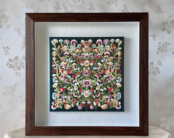 May include: A framed embroidery piece with a dark green background and a floral design in shades of pink, white, and green. The embroidery is surrounded by a white mount and a dark brown frame.