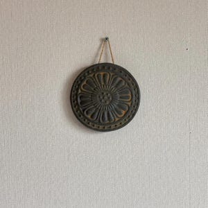 May include: A round, dark metal wall hanging with a floral design. The decorative piece is suspended by a natural fiber cord from a screw in the wall. The metal has a distressed, aged appearance with a central flower and dotted border.