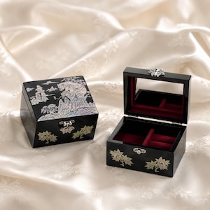 May include: Two black lacquer jewellery boxes with mother-of-pearl inlay. One box is closed, showing a mountain landscape, while the other is open, revealing a red velvet interior, a mirror, and a silver butterfly latch on the lid.