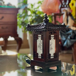 Könnte beinhalten: Eine Tischlampe aus dunklem Holz mit traditionellem asiatischem Design. Die Lampe hat einen quadratischen Sockel und ein Dach im Pagodenstil. Der Lampenschirm ist aus weißem Papier mit einem Blumenmuster.