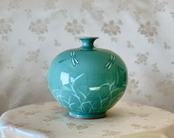 May include: A round, light blue ceramic vase with a dragonfly design. The vase has a textured surface and a glossy finish.