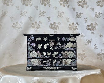 Pode incluir: Um porta-joias preto com quatro gavetas, adornado com desenhos florais e de borboletas em madrepérola. O porta-joias está sobre uma toalha de mesa branca, com um fundo floral. O porta-joias tem um topo curvo e pés decorativos.