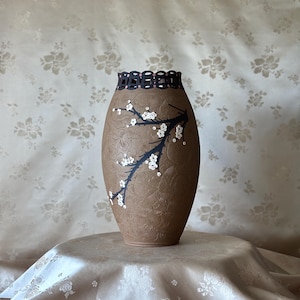 May include: A brown ceramic vase with a textured floral pattern across the surface. A black branch design is painted on the front, adorned with small white flower-shaped sequins. The rim features a dark, intricate, openwork cutout border.