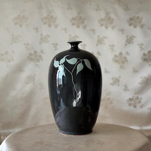 May include: A glossy black ceramic vase with a narrow neck and a rounded body. The front of the vase features a hand-painted design of a delicate, thin-stemmed plant with light mint green leaves, creating a contrast against the dark surface.