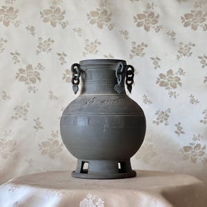 Può includere: Un vaso di ceramica grigia con corpo bulboso e base svasata. Il vaso presenta due manici con maglie decorative a catena e figure di animali incise intorno al centro. Il vaso è esposto su un tavolo con una tovaglia a motivi floreali.