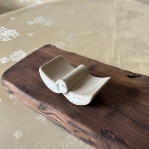 May include: A beige ceramic spoon rest with a curved design, resting on a dark wooden cutting board. The spoon rest has a small, cylindrical handle and delicate floral accents. The cutting board has a natural, live edge.