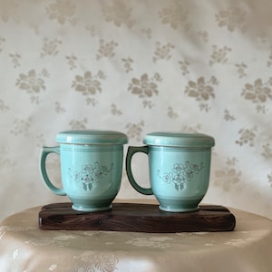 May include: Two matching light teal ceramic mugs with lids, each featuring a floral design and a handle. The mugs are displayed on a dark wooden tray. The background has a floral pattern.