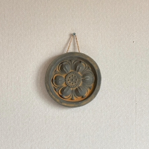May include: A round, gray wall hanging with a floral design. The flower has a detailed center and petals, with a contrasting orange-brown color. It is suspended by a natural fiber cord from a screw in the wall.