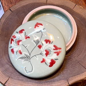 May include: A round ceramic lid with a floral design. The lid is painted with white and red flowers on a light green background.
