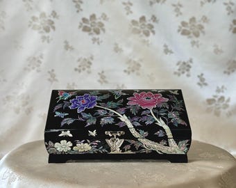 Pode incluir: Uma caixa de joias de madeira preta com uma tampa articulada. A caixa é decorada com um desenho floral feito de incrustações de madrepérola. As flores são rosa, roxas e brancas. Também há borboletas no desenho.