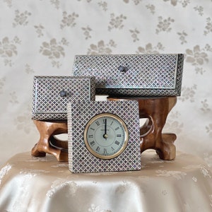 May include: Three decorative boxes made of mother of pearl with a geometric pattern. The boxes are white with iridescent purple and blue accents. The largest box is rectangular and the two smaller boxes are square. The smaller square box is a clock with Roman numerals and a gold face.