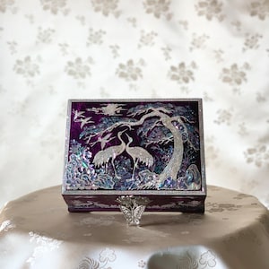 May include: A purple wooden box with a hinged lid decorated with a mother-of-pearl inlay of two cranes standing in a pine tree. The box has a silver butterfly-shaped clasp.