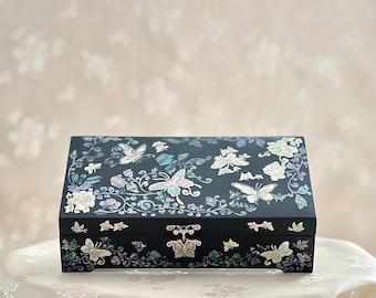 Op de afbeelding: Een zwarte houten doos met een scharnierend deksel, versierd met een bloemen- en vlinderontwerp gemaakt van parelmoer. De doos heeft een zilveren sluiting.