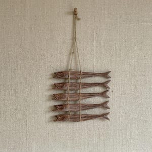 May include: A decorative wall hanging featuring five wooden fish, each with detailed carvings. The fish are strung together with twine and suspended from a wooden peg. The fish are a natural brown color.