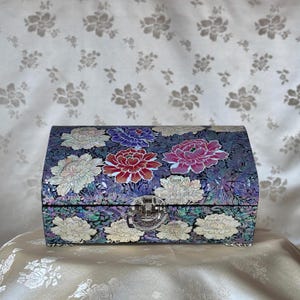 Puede incluir: Un joyero decorativo con tapa curva, adornado con un estampado floral. La caja presenta un fondo oscuro con flores grandes y coloridas en tonos rojos, morados y blancos. Un cierre plateado asegura la caja.