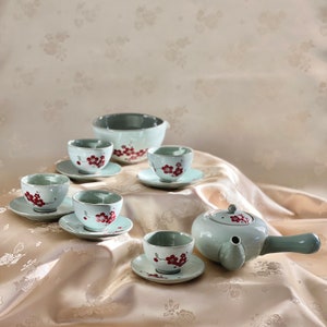 May include: A light blue tea set with a teapot, a bowl, and five cups. The cups and teapot have a floral design with red flowers. The set is on a light brown surface.