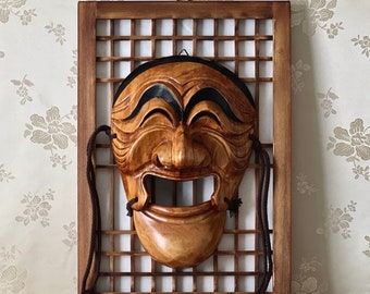 May include: A carved wooden mask with a polished light brown finish, featuring a wide open mouth, prominent nose, and black painted eyebrows. The mask is mounted on a rectangular wooden grid frame with a dark brown finish and hanging cords.