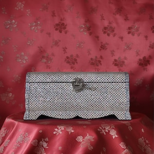 Pode incluir: Um baú decorativo retangular com exterior estampado e fecho de metal. O baú está sobre uma toalha de mesa vermelha com um desenho floral, contra um fundo floral vermelho. O design do baú apresenta um padrão geométrico repetitivo.