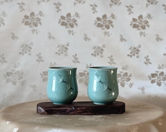May include: Two light blue ceramic teacups with a white crane design on a dark brown wooden tray. The cups are sitting on a light beige fabric surface.