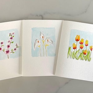 May include: Three blank greeting cards with watercolor floral designs. One card features a branch with pink blossoms, another shows calla lilies, and the third displays yellow and orange tulips. Each design is set against a light blue square.