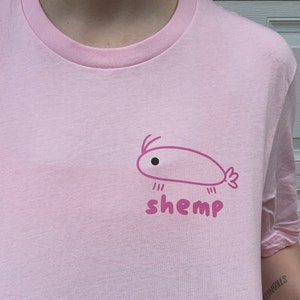 Könnte beinhalten: Hellrosa T-Shirt mit einem kleinen Cartoon-Garnelen-Motiv und dem Wort "shemp" in Dunkelrosa. Das Shirt hat einen Rundhalsausschnitt und kurze Ärmel.