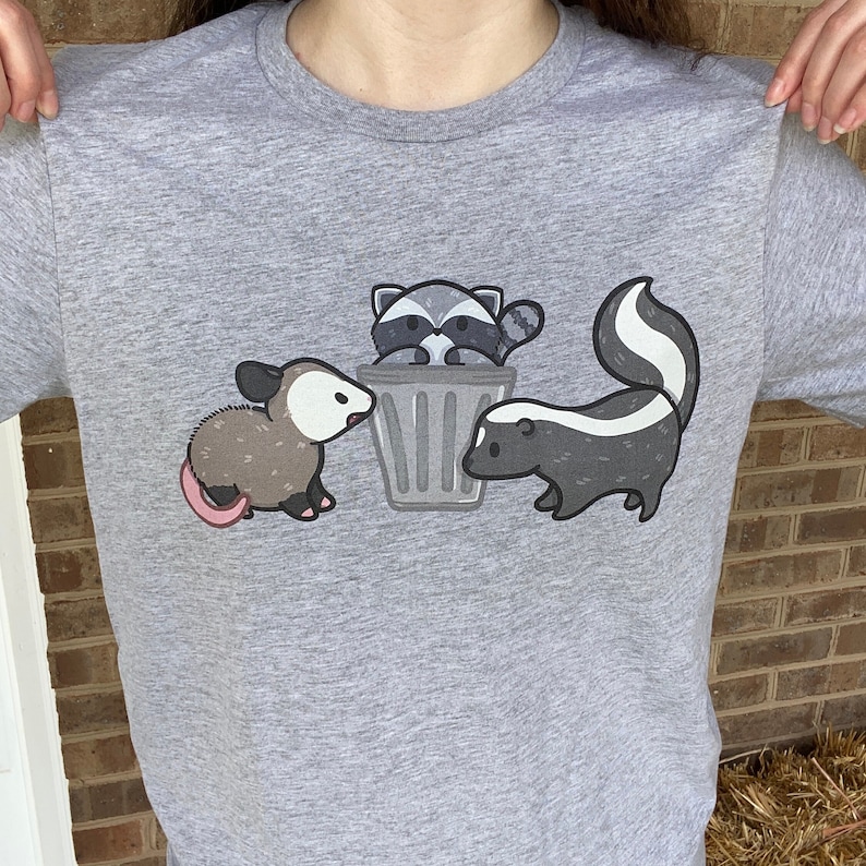 May include: A gray t-shirt with a cartoon illustration of a raccoon in a trash can, a skunk sniffing the trash can, and an opossum looking at the trash can.