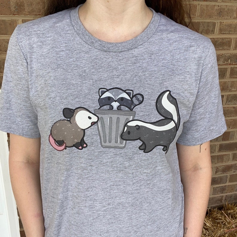 May include: A gray t-shirt with a graphic of a raccoon, skunk, and opossum. The raccoon is in a trash can, the skunk is sniffing the trash can, and the opossum is looking at the skunk.