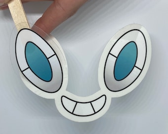 May include: A white sticker with a cartoon face design. The design features two oval-shaped eyes with blue centers and a smiling mouth. The sticker is held by a wooden stick.