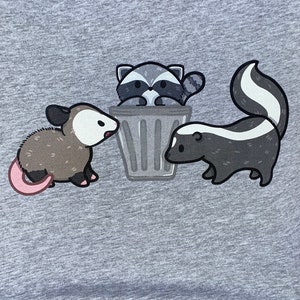 May include: A gray t-shirt with a cartoon illustration of a raccoon in a trash can, a skunk, and an opossum.