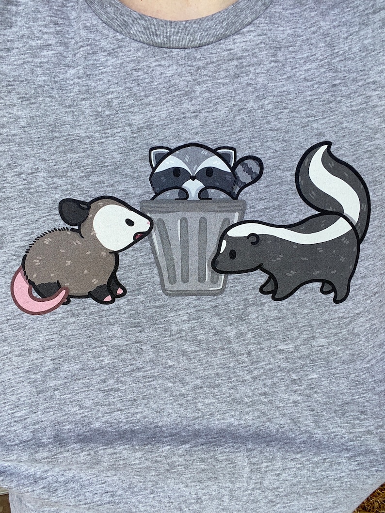 May include: A gray t-shirt with a cartoon illustration of a raccoon in a trash can, a skunk, and an opossum.