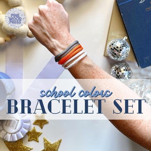 May include: A set of school colors bracelets in gray, orange, and white. The bracelets are displayed on a person's arm. The text "school colors BRACELET SET" is overlaid on the image. The Pine Wood Press logo is in the upper left corner.