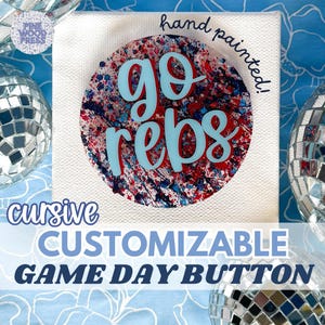 May include: A white canvas with a colorful, hand-painted design. The design features a red, white, and blue splatter-paint effect within a circle, with the words "go rebs" in light blue cursive. The text "CUSTOMIZABLE GAME DAY BUTTON" is below.